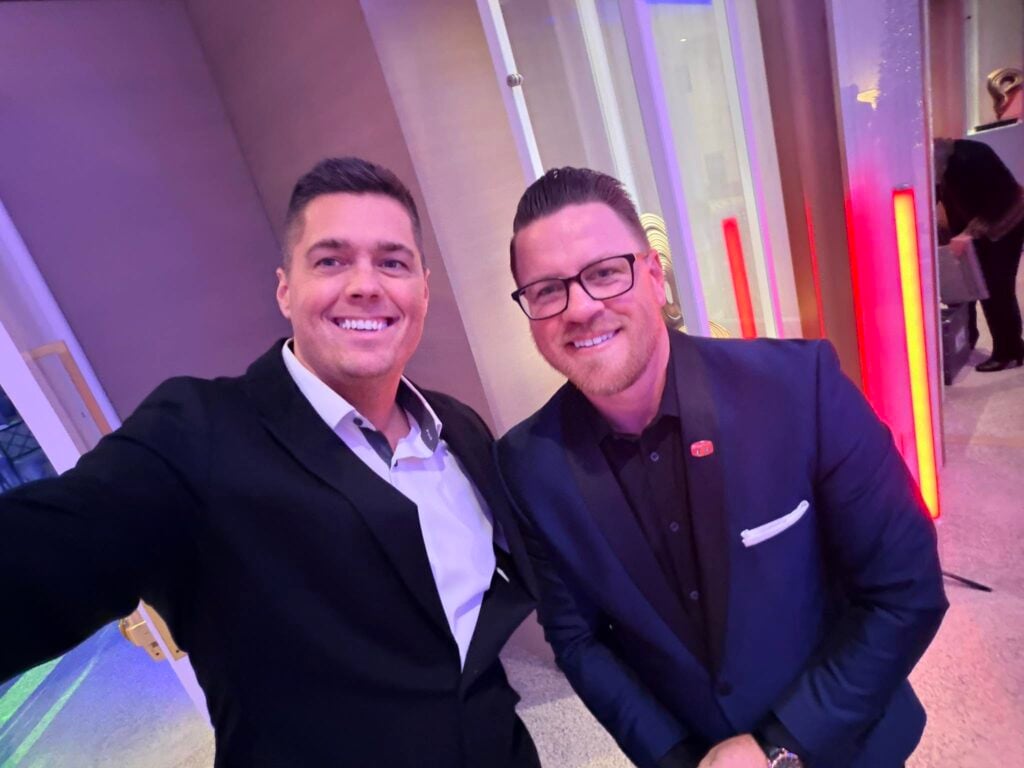 Two leading plumbing influencers in suits take a selfie indoors at Hook Agency, with modern lighting and red and yellow vertical illuminated panels in the background.