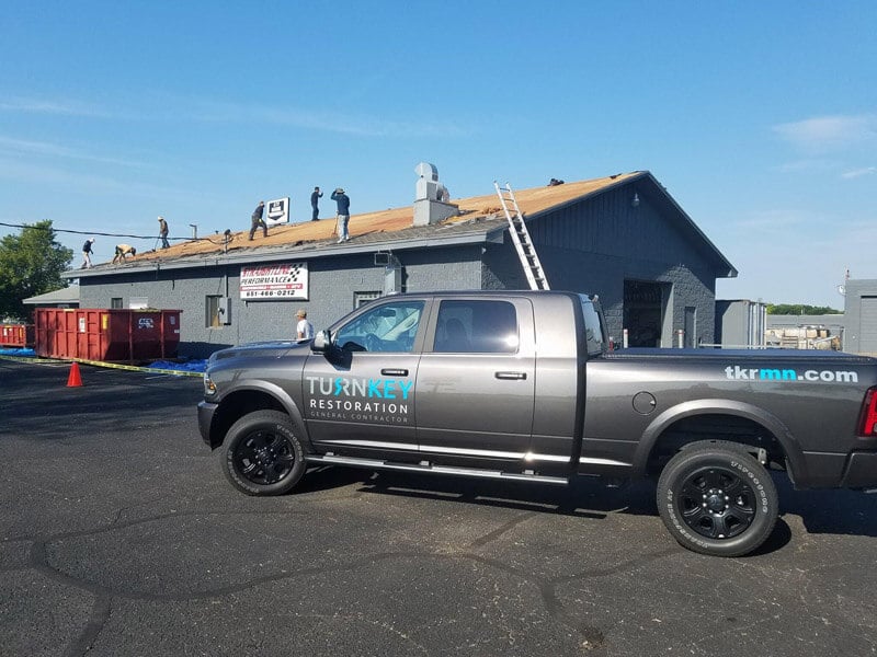 A gray pickup truck featuring "Turnkey Restoration" branding is parked in front of a building undergoing residential roof repairs by U.S. contractors. Ladders are positioned against the structure, and a red dumpster is onsite for debris removal.