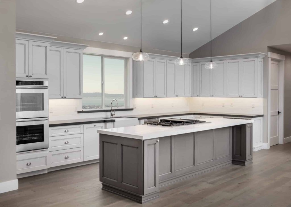 The modern kitchen at Hook Agency showcases sleek white cabinets, stainless steel double ovens, and a spacious gray island with an integrated stovetop. Pendant lights illuminate the island, while a window frames picturesque views of water and distant landscapes. Light wood flooring adds the finishing touch to this elegant design.