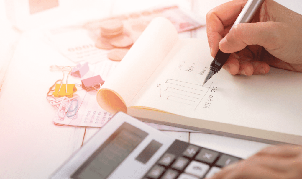 A person writing on a notepad with a pen, next to a calculator, breaks down the marketing budget. The table is cluttered with paper, clips, and coins, suggesting a financial theme involving calculations or budgeting.