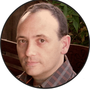 A person with short dark hair, wearing a checkered shirt, looks directly at the camera. The background features green foliage and partially visible wooden details, capturing one of the best parts of working full-time freelancing: enjoying nature as your office.