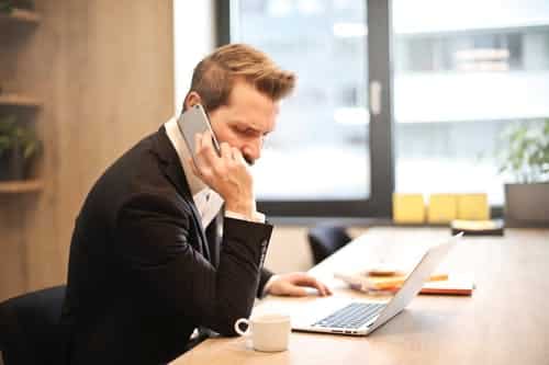 On Hook Agency's website, a professional in formal attire is depicted strategizing lead generation. He's engaged in a smartphone conversation while simultaneously working on his laptop. A small cup is placed prominently, and the background reveals a building view through the windows.