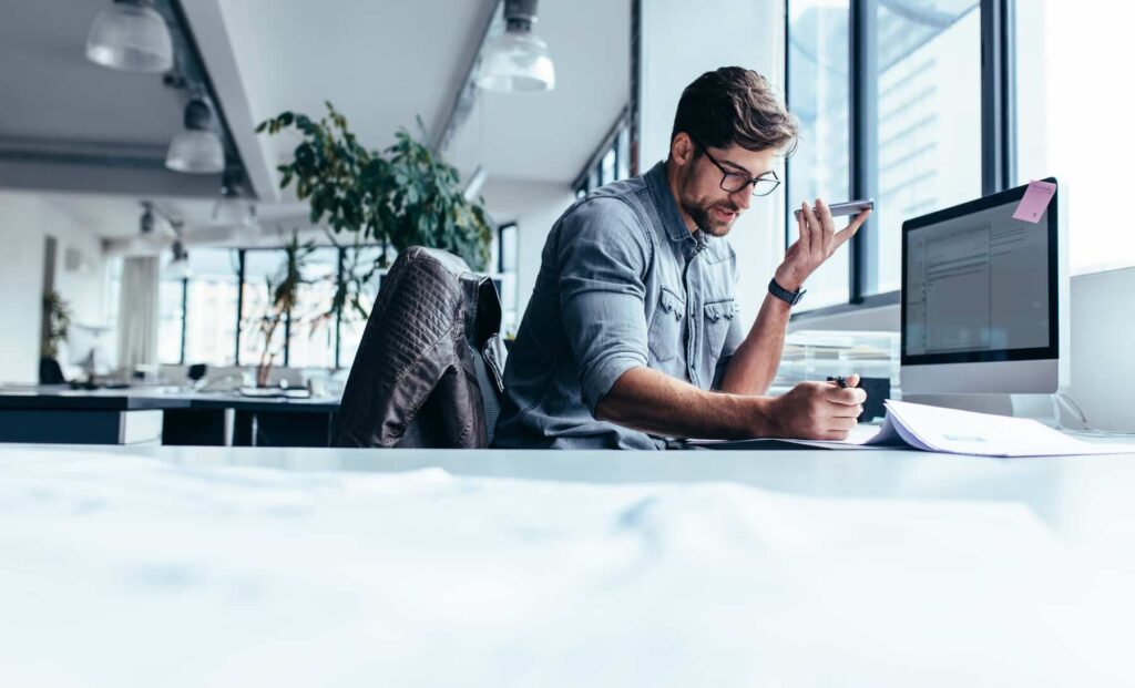 In a modern office, a professional focuses on niche provider strategies, speaking into his smartphone with pen in hand. He is deeply engaged in keyword research at his cluttered desk, illuminated by large windows and a jacket casually draped over his chair.