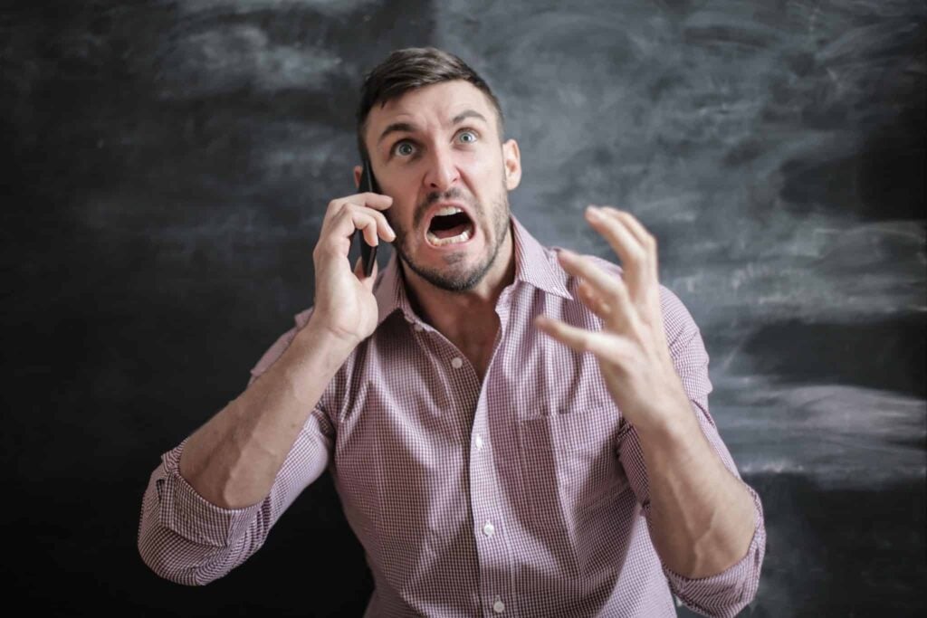 A man in a checkered shirt stands before a blackboard, angrily speaking on his cellphone. His eyes are wide, mouth open in frustration, and his left hand is raised as he addresses pressing sales issues.