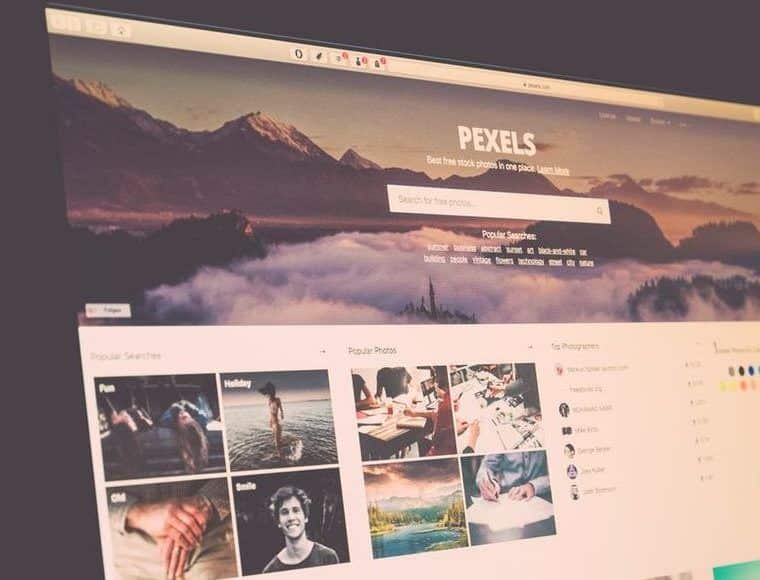 A computer screen displays the Pexels website homepage, designed to help users avoid copyright infringement. The layout includes a search bar, popular searches like "fun" and "holiday," and a grid of photos featuring landscapes and people. The background image shows a misty mountain scene under a cloudy sky.