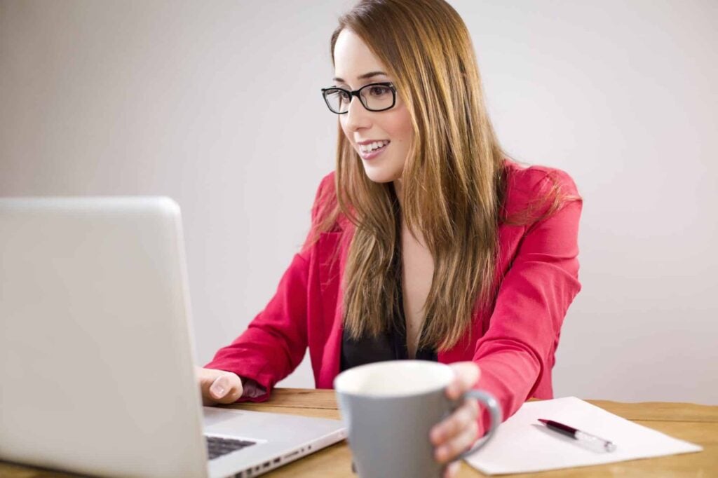 A professional woman in a red blazer, wearing glasses, confidently smiles as she works on her laptop.