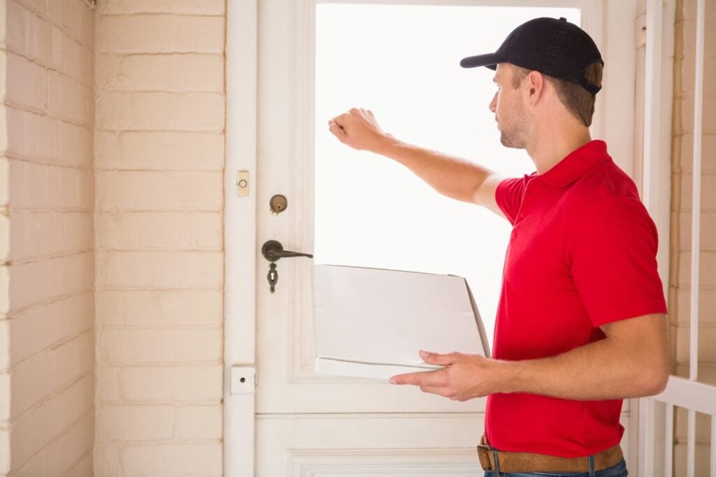 A delivery person in a red shirt and cap knocks on a white door, package in hand. The bright daylight illuminates the porch, set against a brick wall. Could this be the key moment for addressing roofing sales objections?