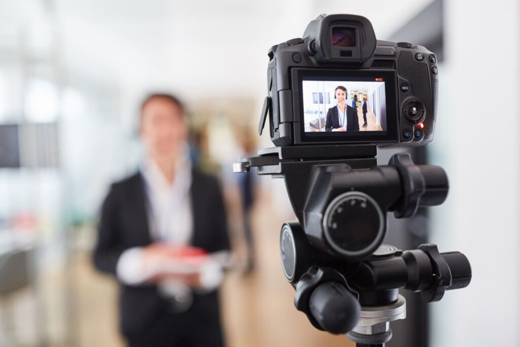 A digital camera mounted on a tripod captures a professional marketing presentation indoors. The screen shows an individual in formal attire, holding a red folder, delivering step-by-step insights. A softly blurred background enhances the professional ambiance.