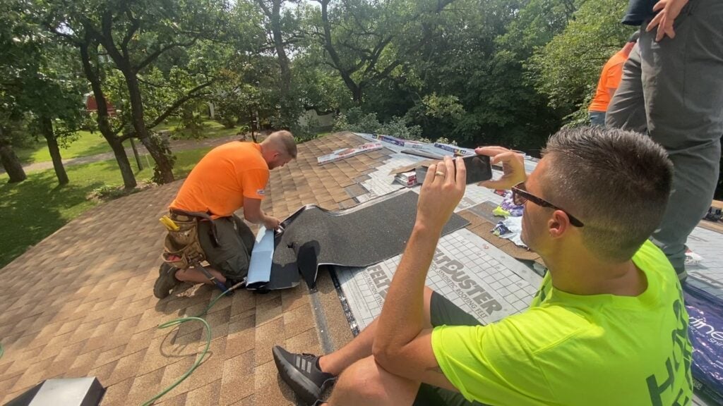 Two men are engaged on a roofing project. The man in an orange shirt installs roofing materials, while the man in a yellow shirt, wearing sunglasses, captures a photo for the Roofing SEO Marketing Guide 2024–2025 cover. Shingles and tools surround them amidst a backdrop of lush greenery.