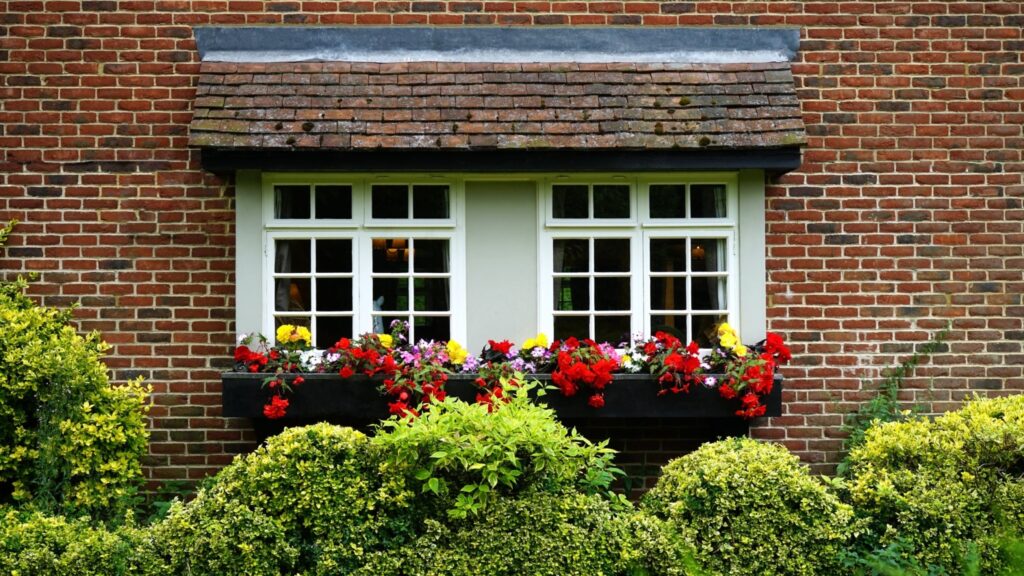 A charming brick house facade features a large window adorned with a wooden flower box brimming with vibrant red, yellow, and purple flowers. Neatly trimmed green bushes below complete this picturesque scene, highlighting the expert design of quality windows and doors.