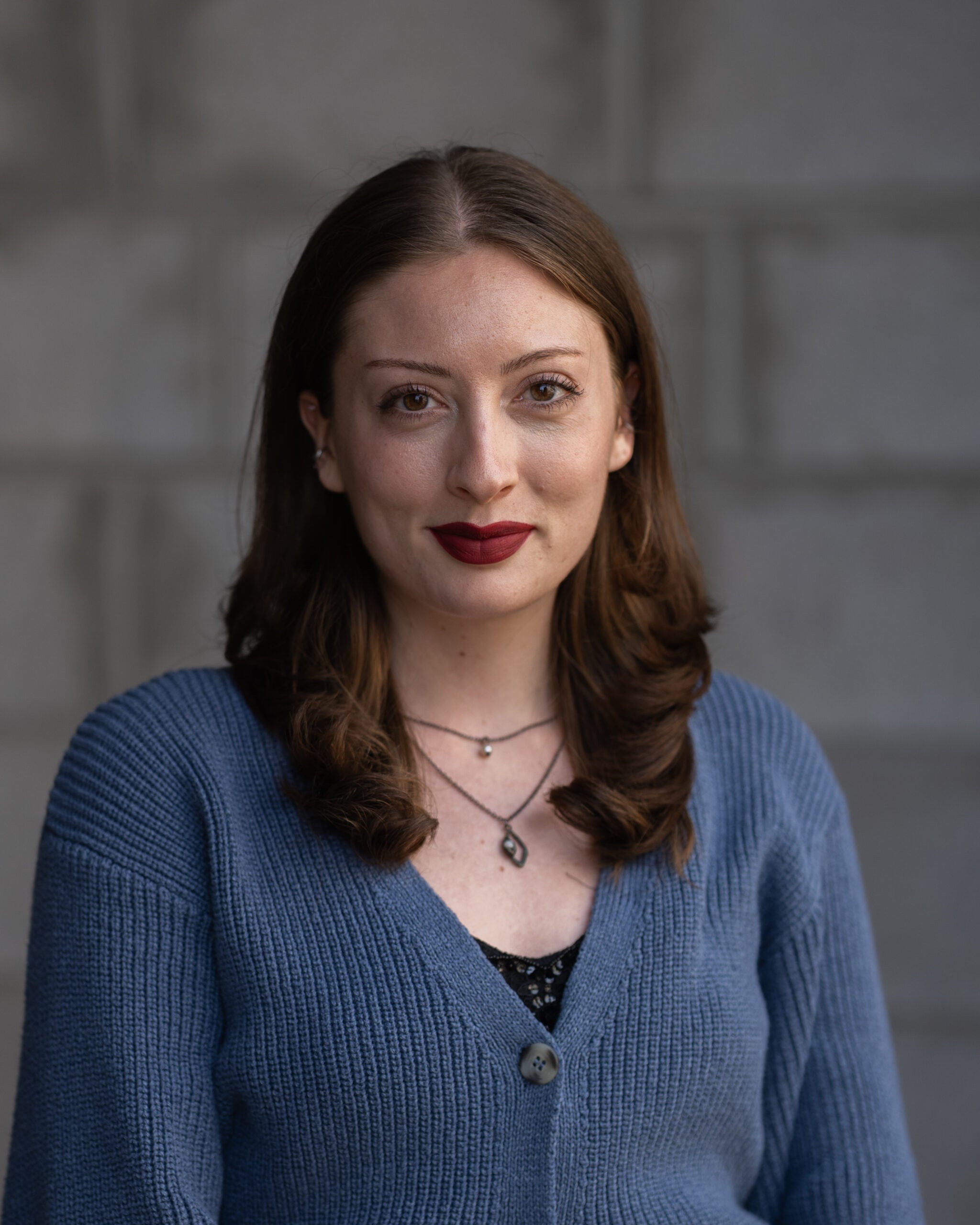 Professional woman with light skin and brown hair in a blue sweater stands before a gray wall, confidently facing the camera.
