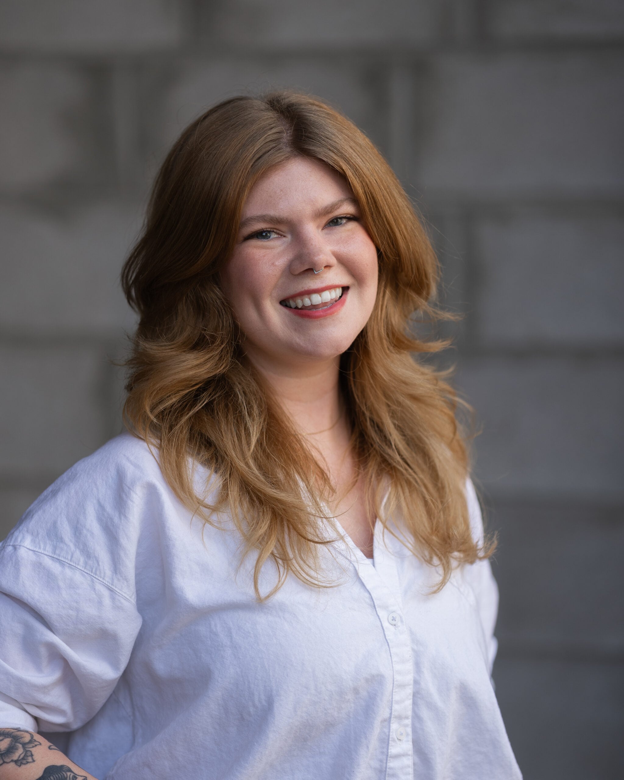 Ashlynn Reynolds, in a white button-up shirt and with long wavy light brown hair, smiles confidently before a gray brick wall.