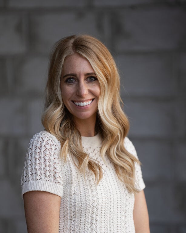 Smiling woman with long, wavy blonde hair in a white knit top stands before a blurred gray brick wall background.
