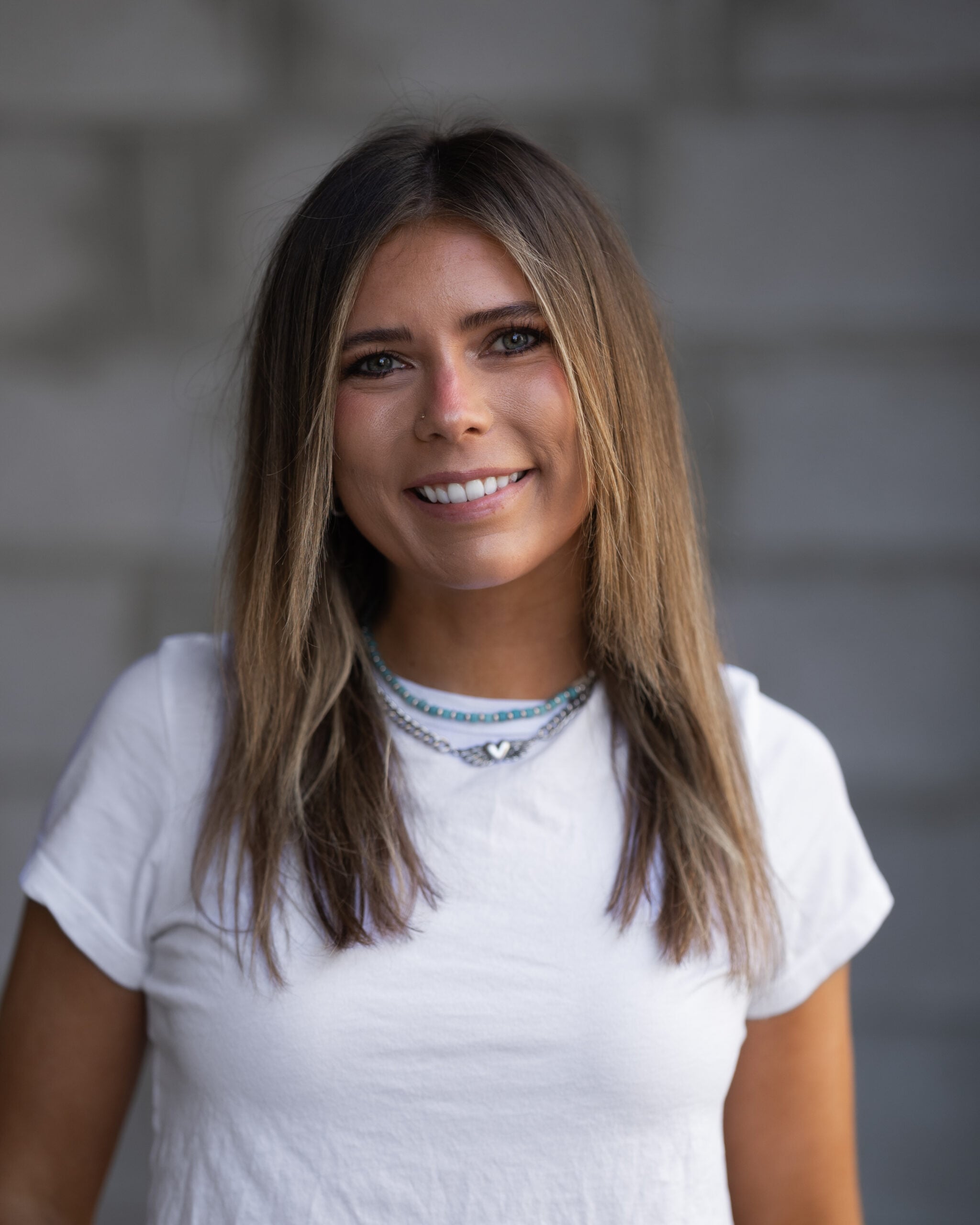 Brynn Wilson, a young woman with straight light brown hair and layered necklaces, smiles in a white t-shirt before a gray brick wall.
