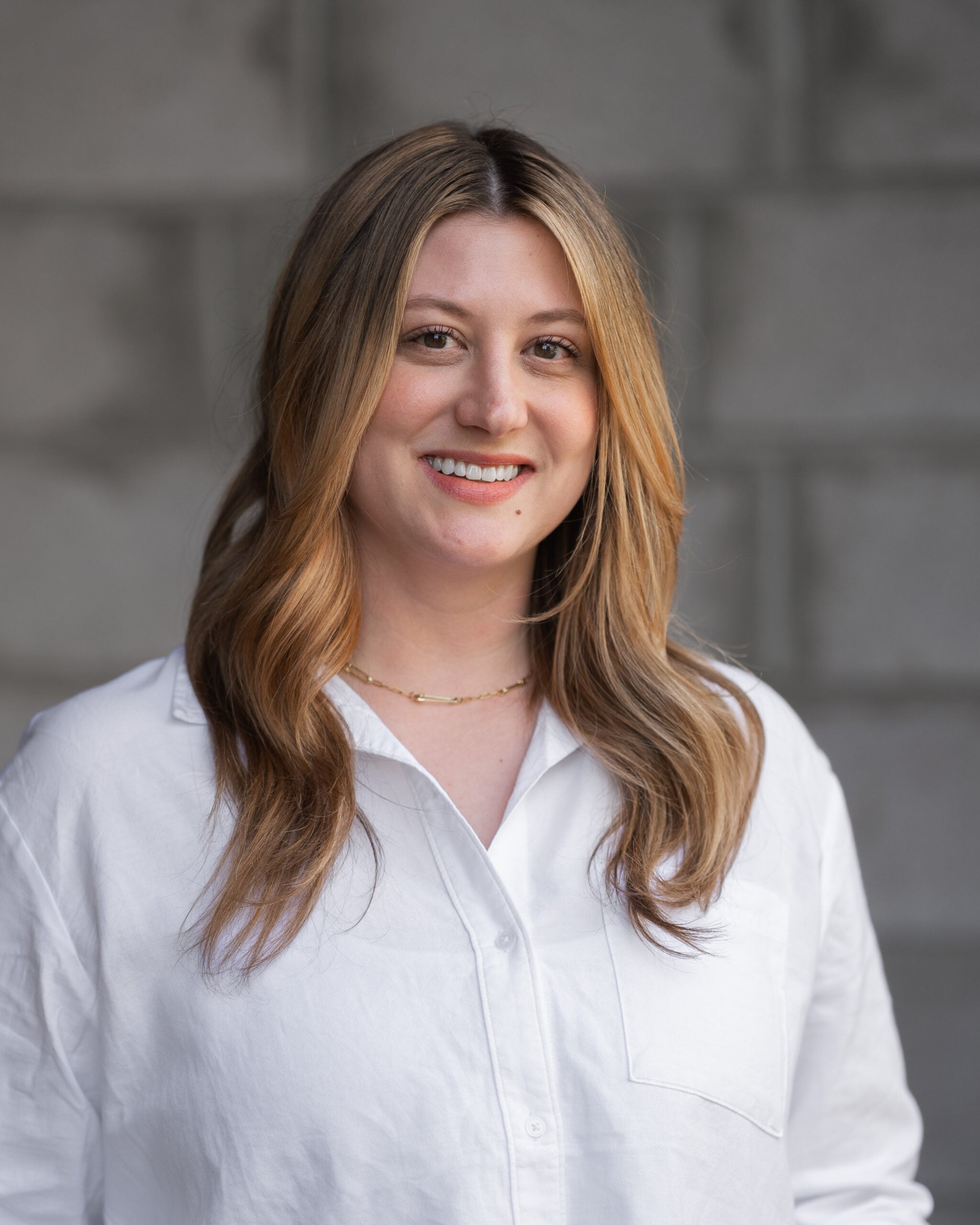 Casey Swanson, with long light brown hair and a white button-up shirt, smiles confidently before a gray brick wall at Hook Agency.