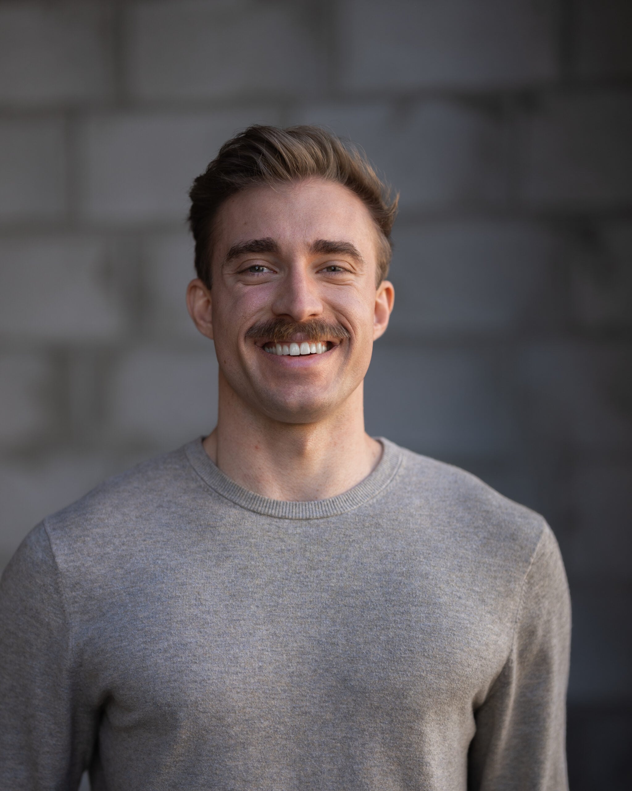Smiling man with short brown hair and mustache, in a light gray sweater, stands before a blurred gray brick wall at Hook Agency.