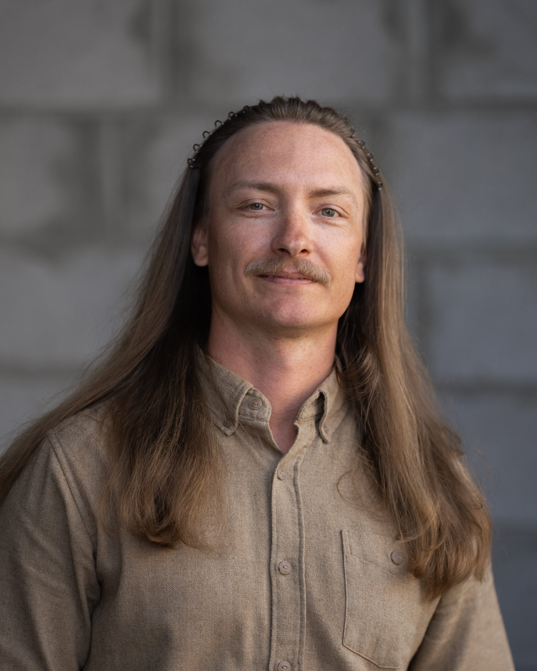 Dillon Treloar stands against a gray stone wall, wearing a beige shirt. He looks directly at the camera with a neutral expression.