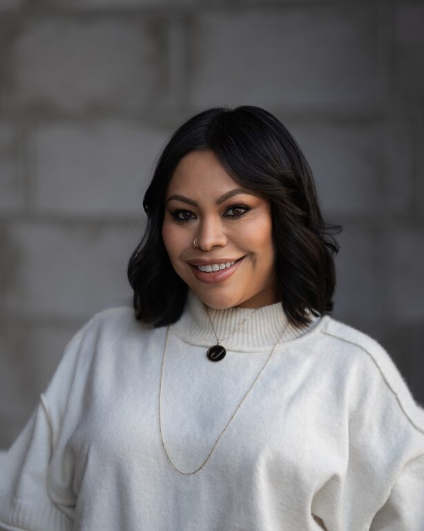 Erin Livingston, with shoulder-length black hair and a cream sweater, smiles confidently at the camera before a gray background.