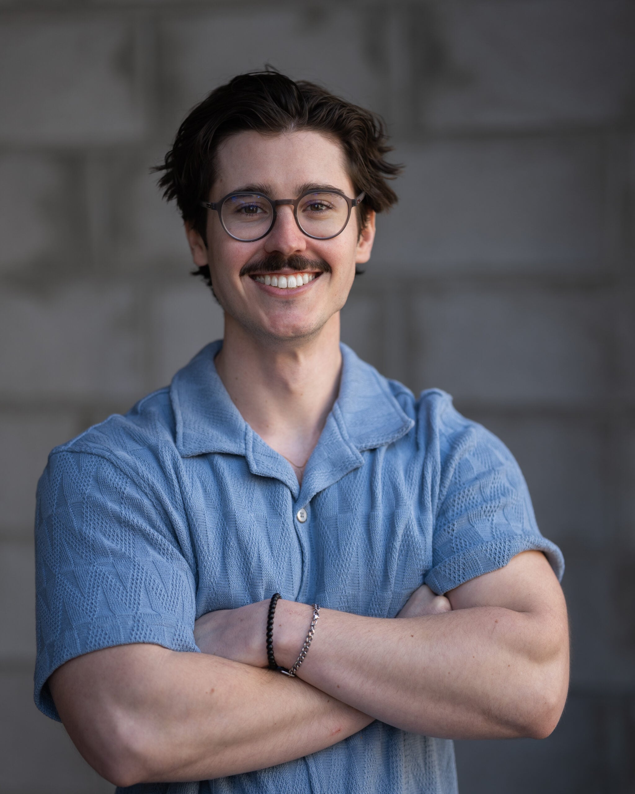 Jameson Kenney, dark hair, glasses, mustache, in light blue short-sleeve shirt, smiles confidently before a gray brick wall.