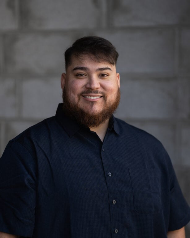 Joel Enrique Palomino, with short dark hair and beard, in a dark button-up shirt, smiling before a gray brick wall at Hook Agency.