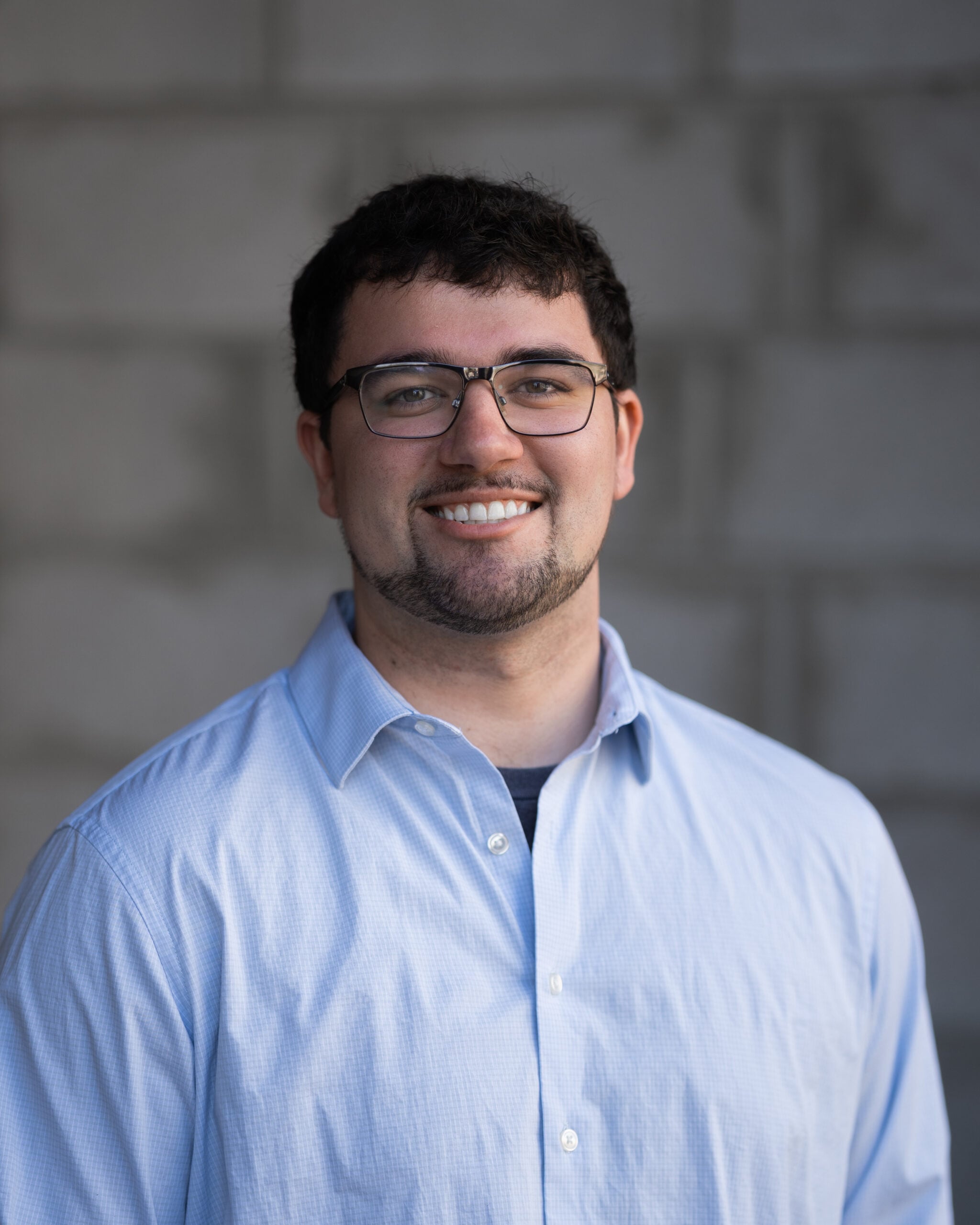 A Hook Agency team member with short dark hair, glasses, and a beard, smiling in a light blue shirt before a gray brick wall.