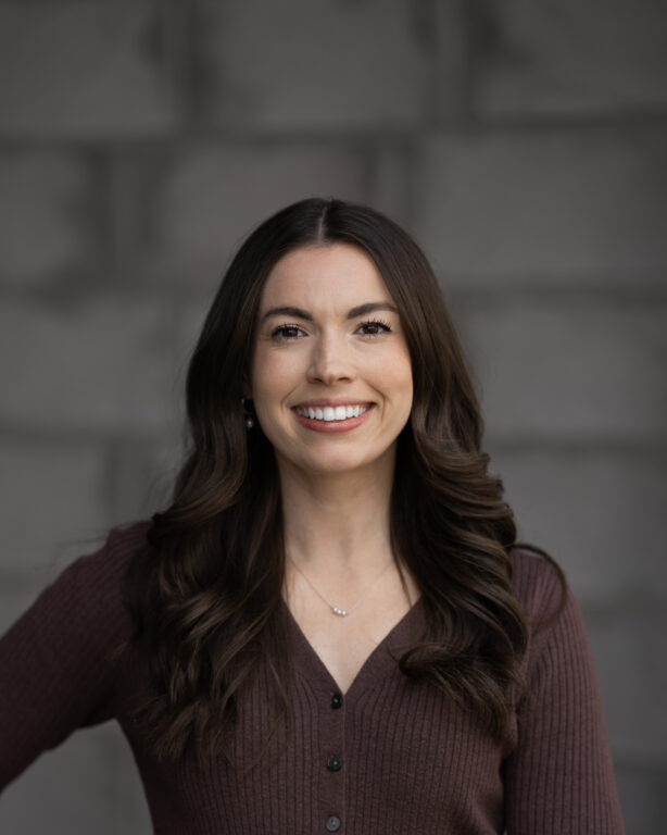 Maggie Benham, with long brown hair and a wide smile, stands in front of a blurred gray stone wall, wearing a brown sweater.