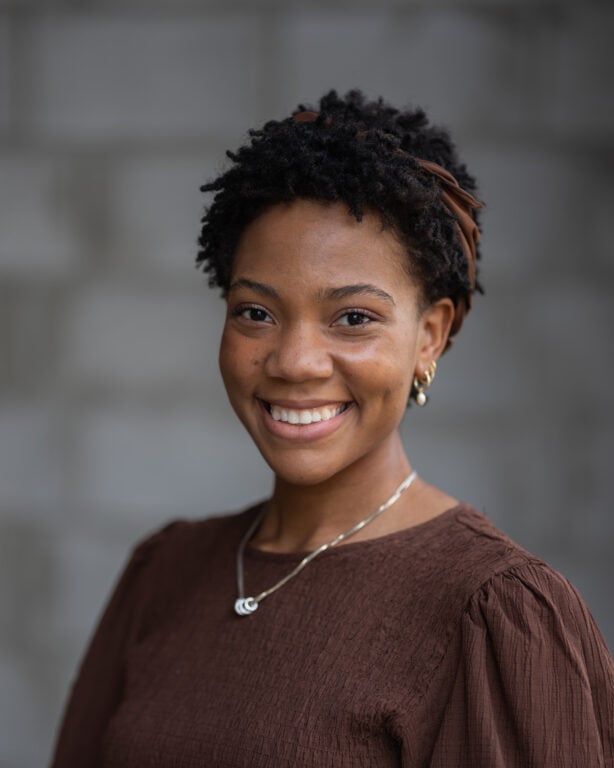 Mandelyn Miller of Hook Agency, with short curly hair and brown headband, smiles in a textured brown top and gold hoop earrings.