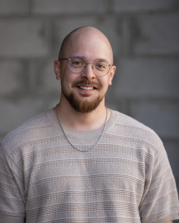 Matt Anderson, smiling, in glasses and a beige sweater, stands before a gray brick wall on Hook Agency’s website.