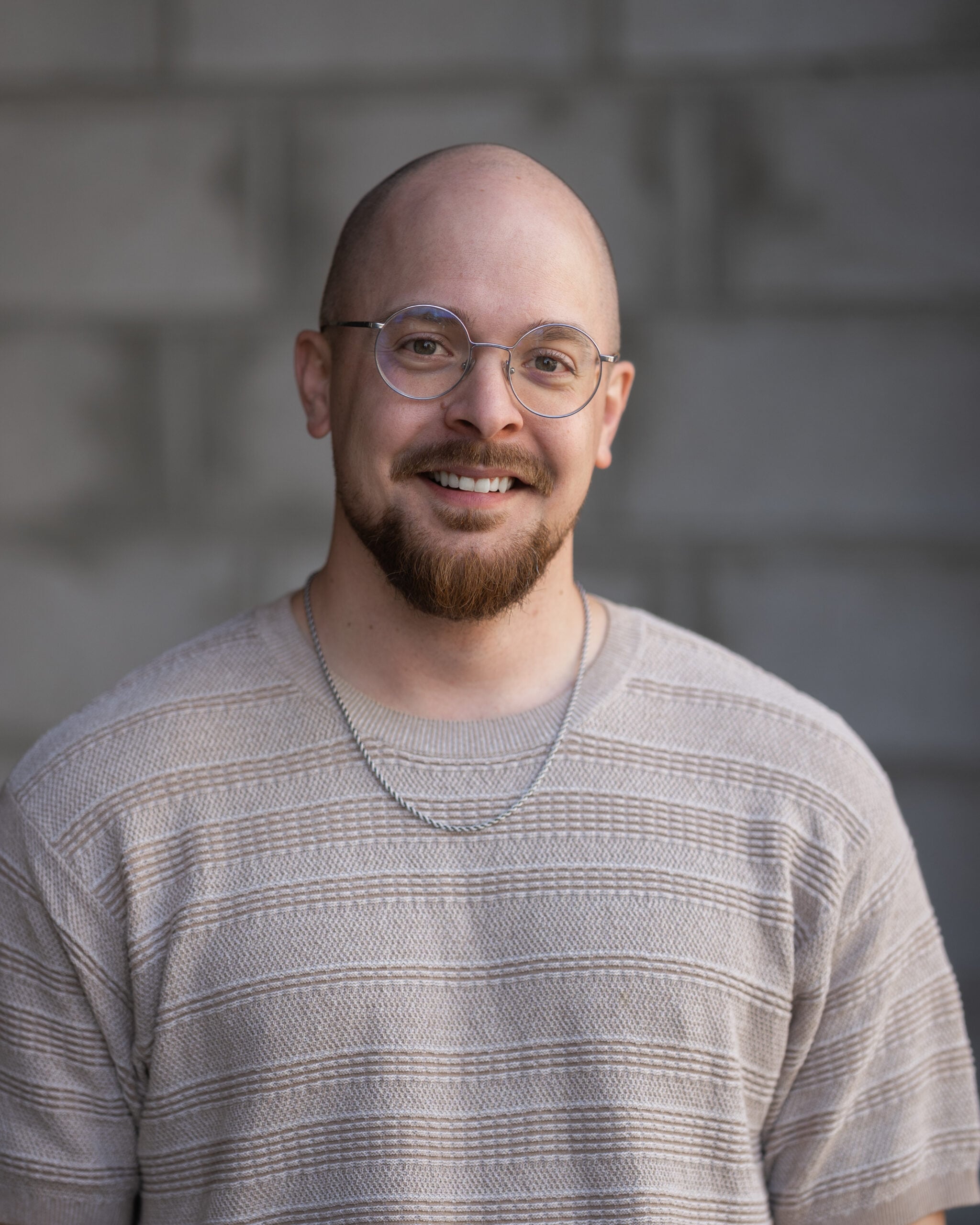 Matt Anderson, smiling, in glasses and a beige sweater, stands before a gray brick wall on Hook Agency’s website.