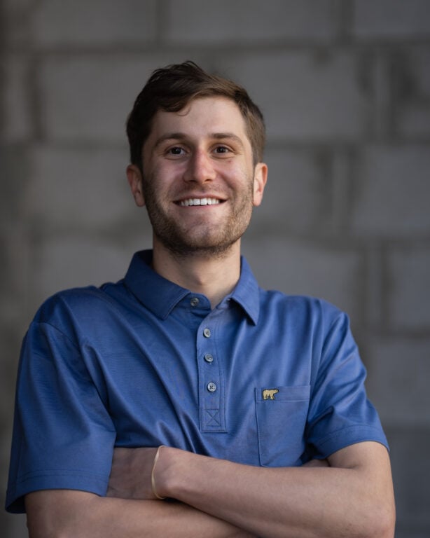 Confident young man with short brown hair in a blue collared shirt stands arms crossed, smiling, in front of a blurred brick wall.