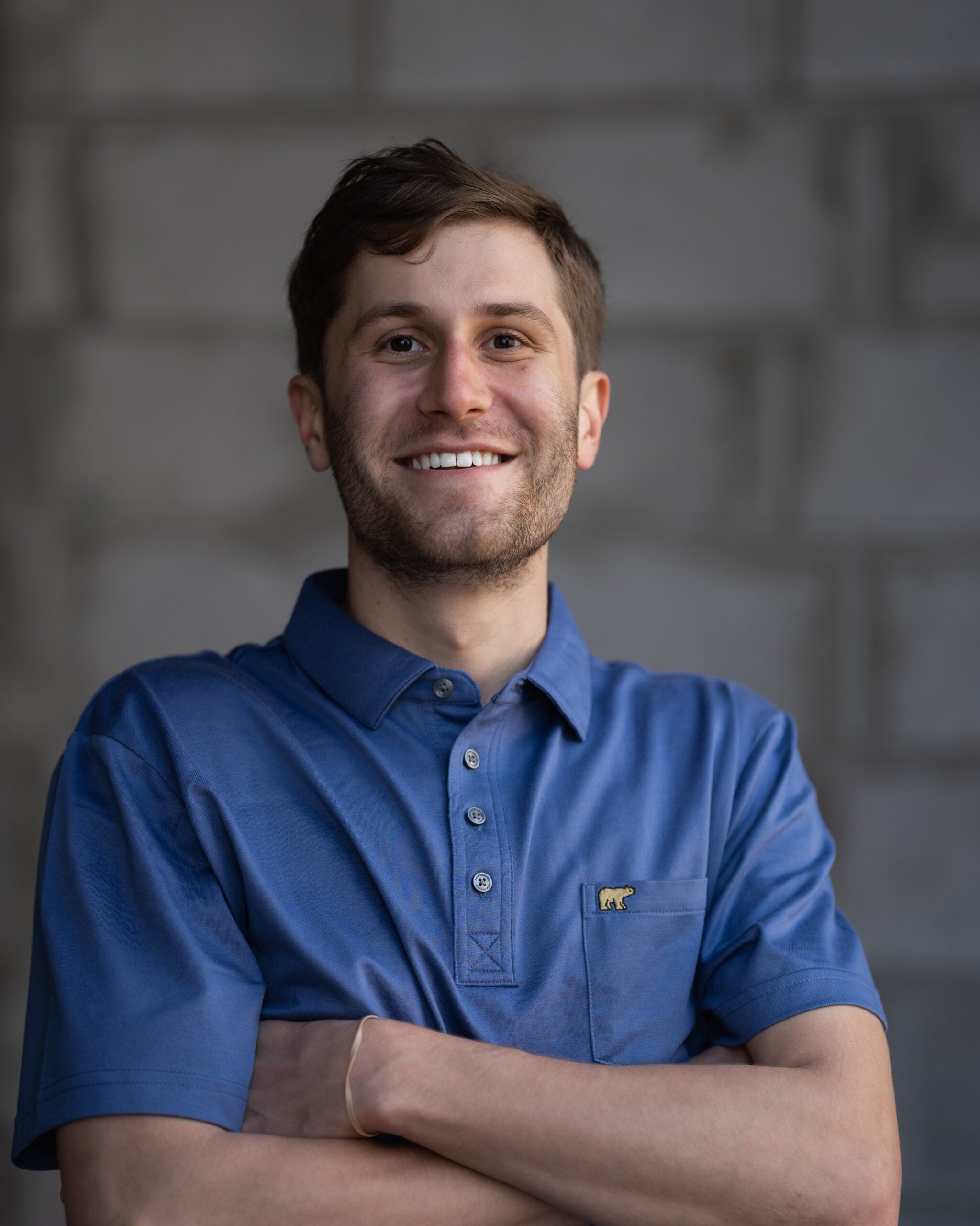 Confident young man with short brown hair in a blue collared shirt stands arms crossed, smiling, in front of a blurred brick wall.