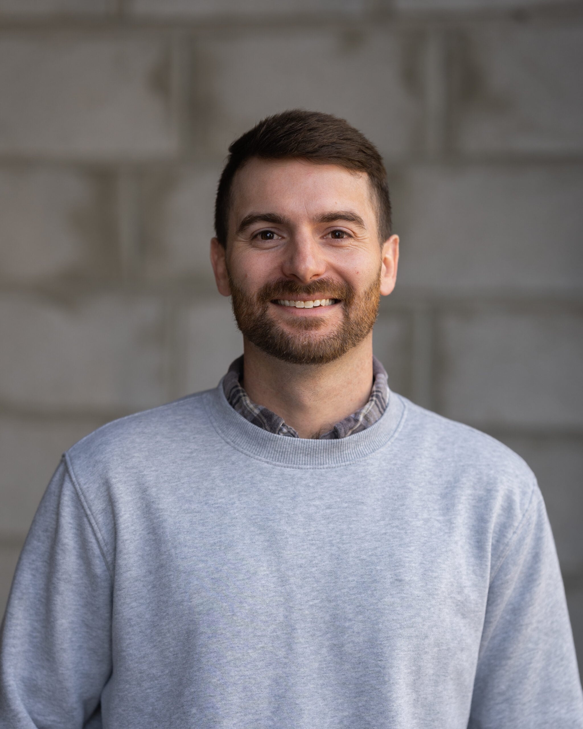 Skyler McDaniel, with short brown hair, beard, and mustache, smiles in a gray sweatshirt over plaid shirt before concrete blocks.
