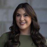 Sydnee Olsen, wearing a green top and with long wavy brown hair, smiles in front of a blurred gray background on Hook Agency’s site.