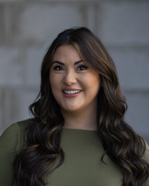 Sydnee Olsen, wearing a green top and with long wavy brown hair, smiles in front of a blurred gray background on Hook Agency’s site.