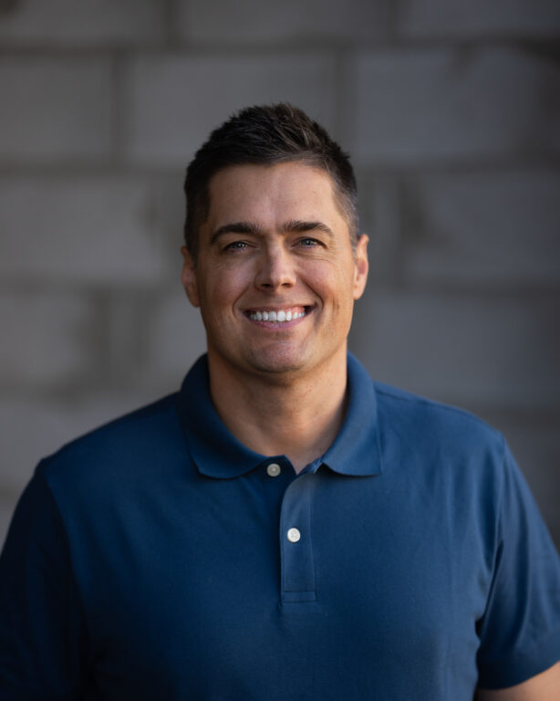 Team member with short dark hair and a blue polo shirt, smiling confidently against a gray brick wall backdrop at Hook Agency.