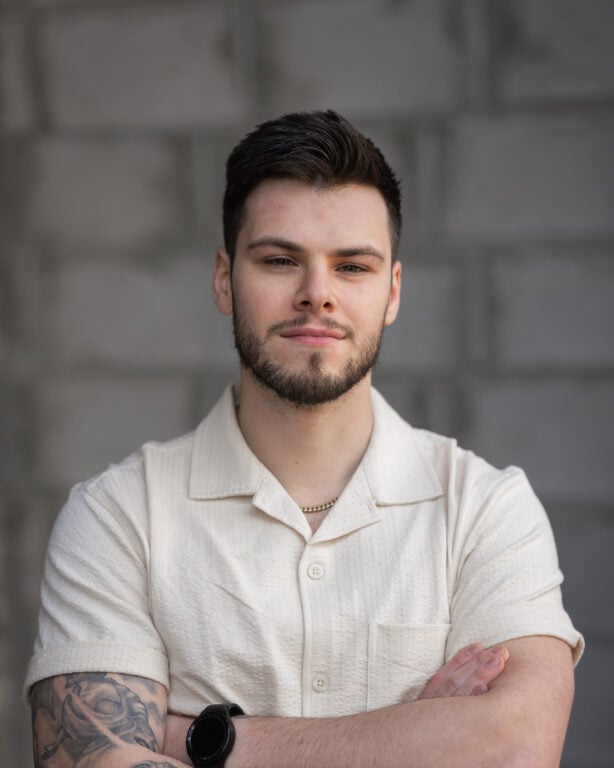 Vincenzo Colosimo stands confidently with arms crossed before a gray brick wall, wearing a light short-sleeve shirt.