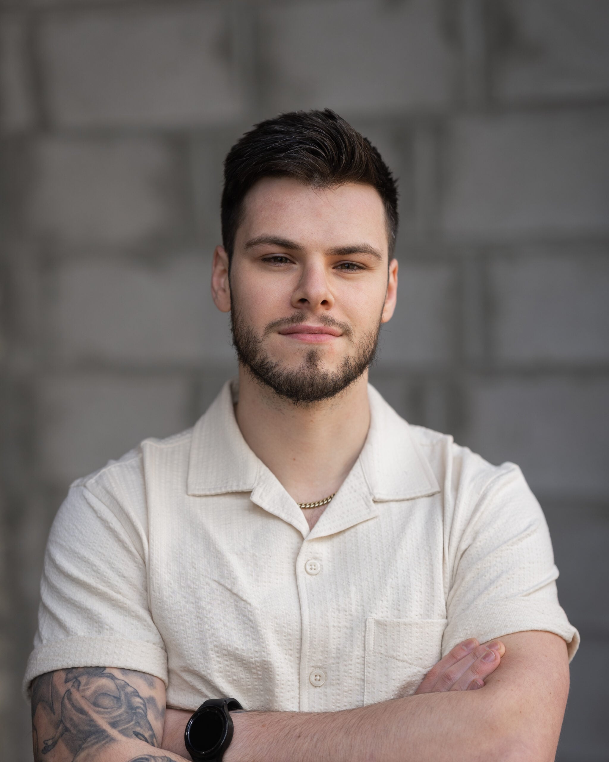Vincenzo Colosimo stands confidently with arms crossed before a gray brick wall, wearing a light short-sleeve shirt.