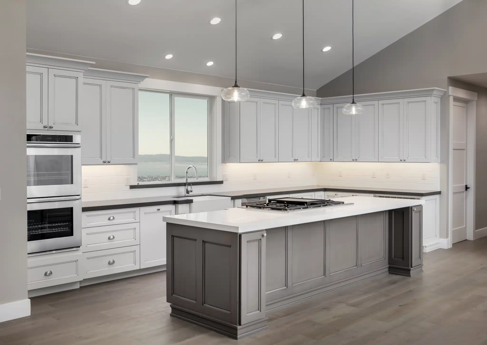 Experience modern kitchen elegance with sleek white cabinets, a sophisticated gray island, and premium stainless steel appliances. Illuminate your space with stylish pendant lights above the island. Enjoy the expansive outdoor view framed like a landscape painting through the large window. Wood flooring beautifully enhances the neutral color palette for a seamless design.