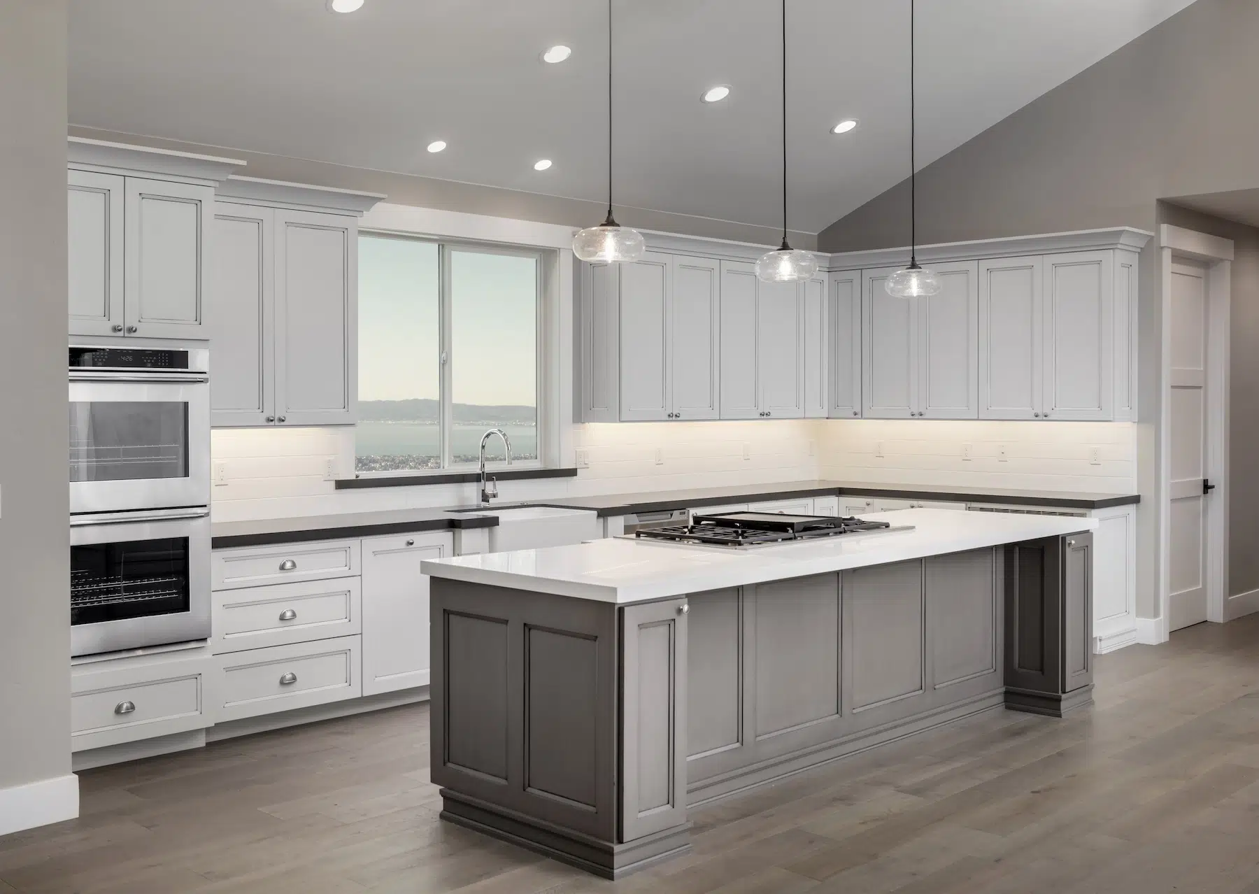 Explore our modern kitchen design featuring sleek white cabinets and a spacious gray island topped with a pristine white countertop. Seamlessly integrated stainless steel appliances, including a double oven, enhance functionality. Elegant pendant lights artfully illuminate the island, complemented by a window view that welcomes the outside world in.