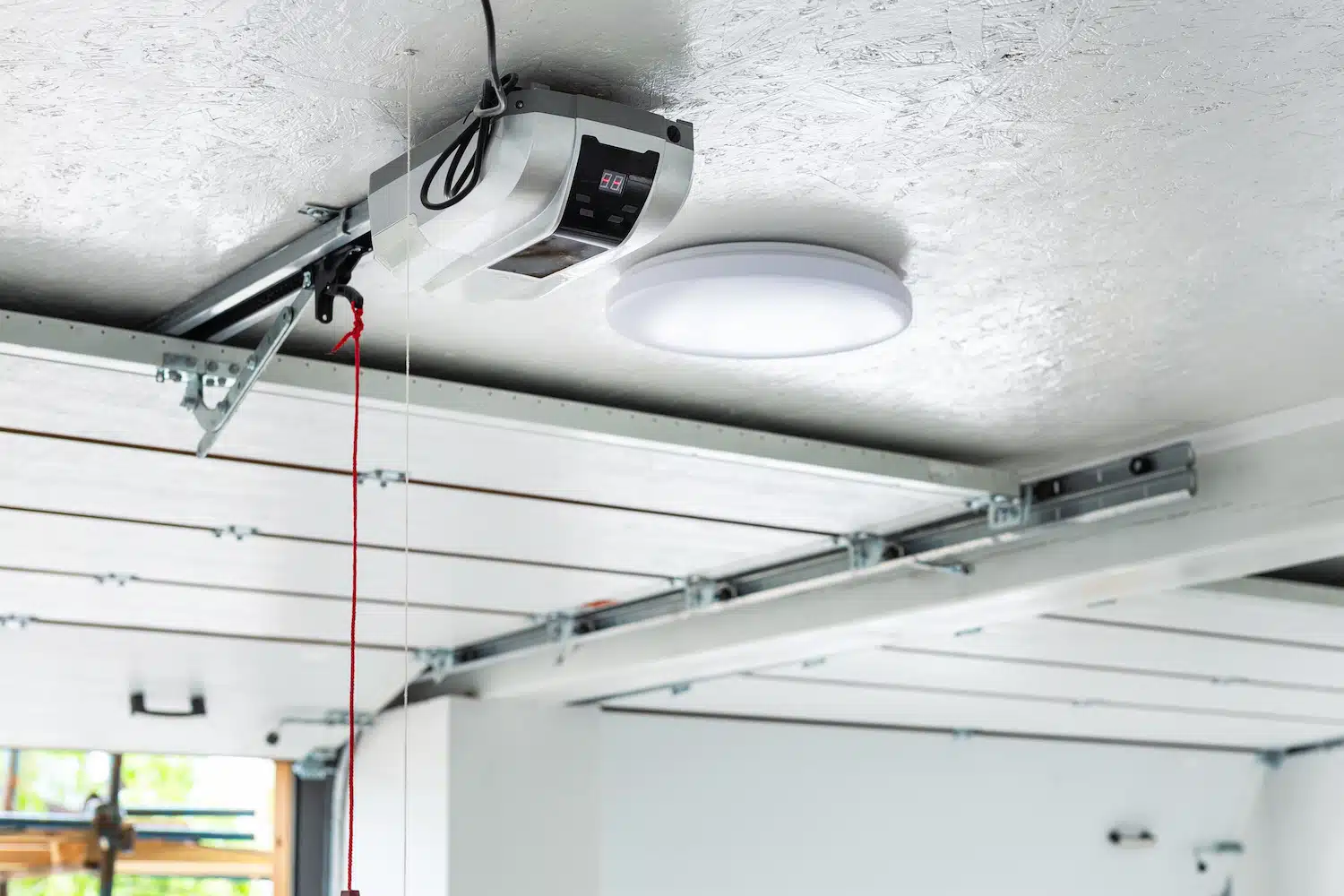 Showcased in the garage is a state-of-the-art automatic garage door opener, accompanied by a circular LED light fixture. The visible tracks ensure seamless door operation, with a red emergency release cord for safety. This setup exemplifies sleek and efficient modern design, ideal for contemporary garage door marketing. best garage door ppc agencies