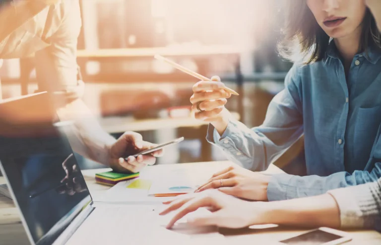 In a dynamic New York office, two professionals collaborate intensely: one focusing on web design details on a laptop, while the other offers insights via smartphone. The workspace is lively with scattered papers and stationery, illuminated by streaming sunlight from the window.