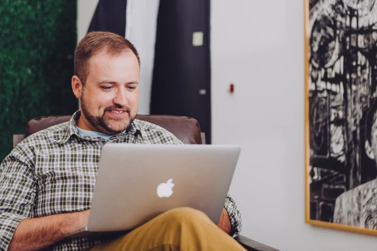 On Hook Agency's website: A man in a plaid shirt and beige pants sits comfortably in a chair, focused on creating the latest San Diego web design. The backdrop features an abstract painting and a green plant, fostering a creative ambiance.