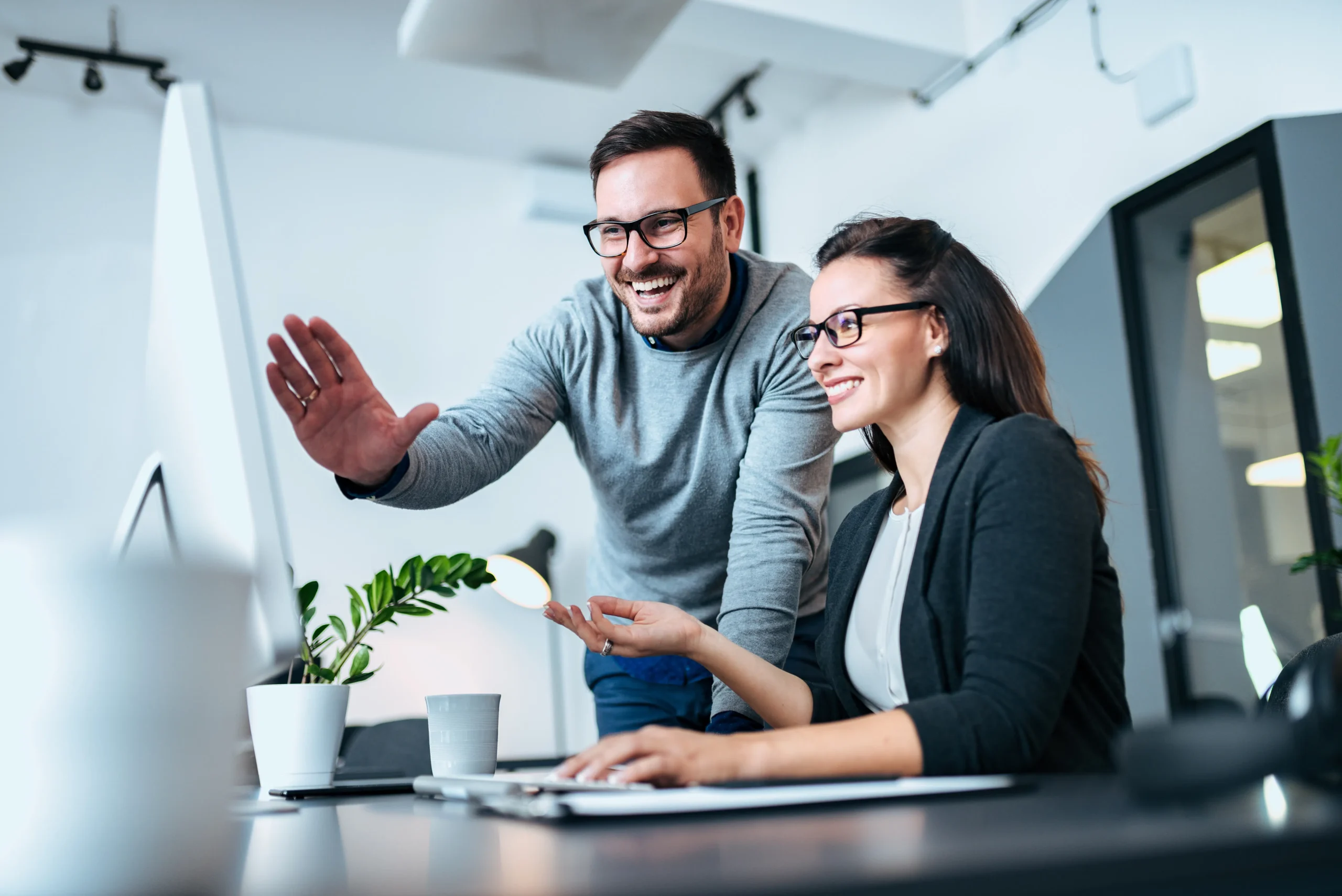 On Hook Agency's Sioux Falls SEO page, a man and woman, both wearing glasses, smile enthusiastically at their computer screen. A small plant and coffee cup rest on the desk as the man gestures with excitement.