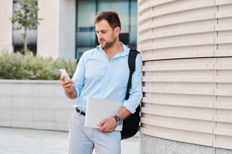 On Hook Agency's website, a professional man wearing a light blue shirt checks the latest Chicago PPC trends on his smartphone. Leaning against an outdoor wall with modern architecture and greenery in the background, he holds a laptop and carries a backpack, adding to his focused and contemporary look.