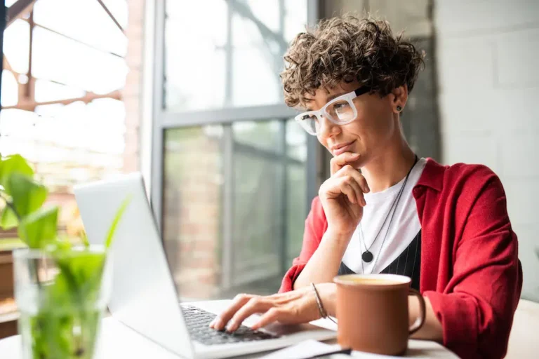On the Hook Agency website, an individual with curly hair and glasses focuses intently on a laptop, symbolizing Chicago web design. Wearing a red shirt, they thoughtfully rest their chin on one hand. The setting includes a mug and small plant on the table, framed by a window in the background.