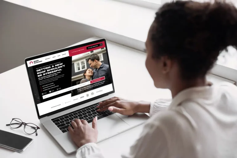 A woman sits at a desk, engaged with her laptop as she navigates a Cincinnati web design site. The display features an image of a smiling couple with accompanying details on roofing services. Nearby, glasses and a notepad are placed neatly on the organized desk.