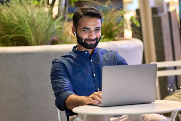 A bearded man in a blue shirt and earbuds focuses intently on his Dallas SEO project, working on a laptop at a small outdoor table surrounded by lush greenery.