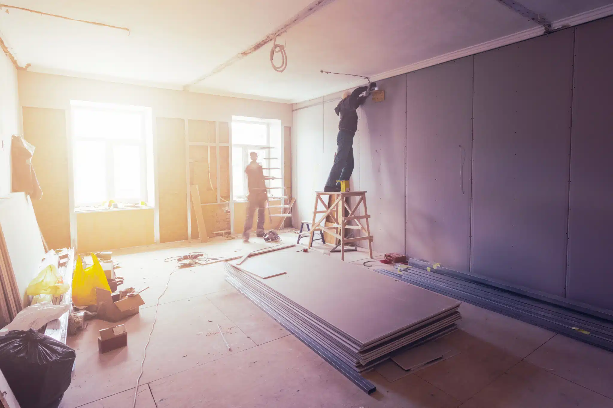 On Hook Agency's website: Two workers actively remodel a room, one positioned on a ladder fine-tuning wall details, while the other is stationed by a window. The environment, teeming with drywall sheets and tools, takes on the appearance of a designer’s canvas. Sunlight pours through the window, emphasizing their skilled craftsmanship.