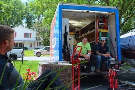 Home Services Marketing Ideas - HVAC SEO Agencies - An HVAC and plumbing truck, emblematic of home service marketing expertise, is stationed on a suburban street. Two individuals sit in its open back, surrounded by tools and a red dolly; nearby, a man relaxes in the yard. The scene is framed by lush trees and a white house with striking red shutters under the clear, sunny sky.