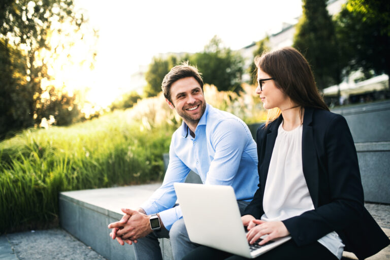A man in a blue shirt and a woman in a black blazer sit together on park steps, discussing Washington DC SEO. She holds a laptop, while he smiles warmly. Lush greenery surrounds them, creating an ideal setting for their conversation.